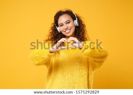 Smiling young african american girl in sweater posing isolated on yellow orange background. People lifestyle concept. Mock up copy space. Listen music with headphones, showing shape heart with hands