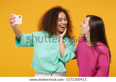 Two women friends european african girls in pink green clothes posing isolated on yellow background. People lifestyle concept. Mock up copy space. Doing selfie shot on mobile phone look at each other