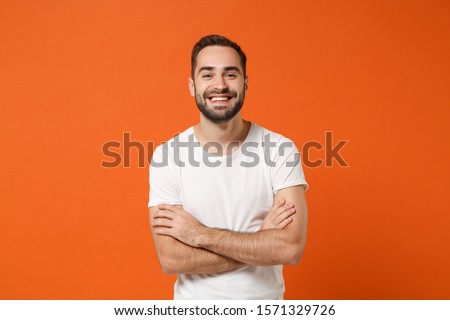 Smiling funny young man in casual white t-shirt posing isolated on bright orange wall background, studio portrait. People sincere emotions lifestyle concept. Mock up copy space. Holding hands crossed