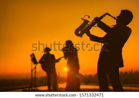 Silhouette autumn or summer scene of saxophone musician man showing with blurry jazz trio band and twilight or sunset city scape background. Image for happy new year party or celebration concept. Royalty-Free Stock Photo #1571332471
