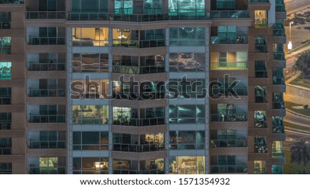 Rows of glowing windows with people in the interior of apartment building at night. Modern skyscraper with glass surface. Traffic on roads on a background. Pan right