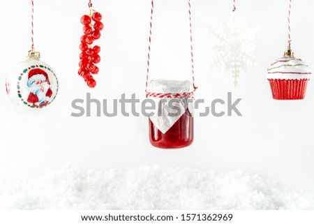Christmas decorations, fresh red currants branch, fruit jam in a glass jar hanging on ropes against white backdrop. Horizontal image with copy space