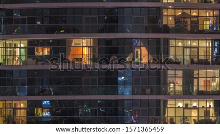 Rows of glowing windows with people in the interior of apartment building at night. Modern skyscraper with glass surface. Concept for business and modern life. Pan right