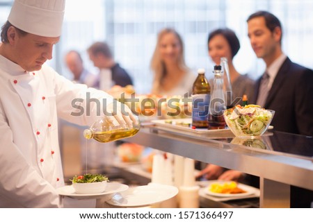 Chef pouring olive oil salad dressing on salad in work cafeteria Royalty-Free Stock Photo #1571366497