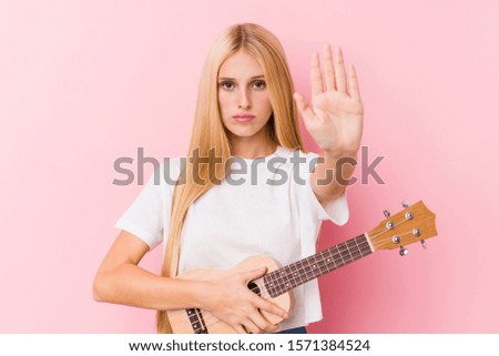 Young blonde girl playing ukelele standing with outstretched hand showing stop sign, preventing you.