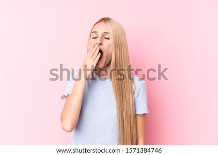 Young blonde woman on pink background yawning showing a tired gesture covering mouth with hand.