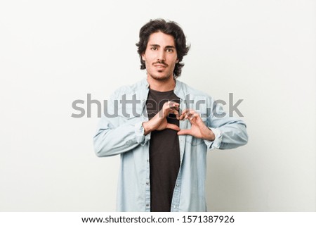 Young handsome man against a white background smiling and showing a heart shape with hands.