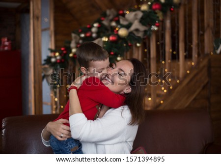 Cheerful and happy mom and little son 4 years old on the background of a wooden house beautifully decorated before Christmas hug each other. A boy kisses mom, and a woman smiles