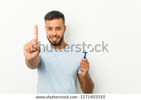 Young south-asian man holding a razor blade showing number one with finger.