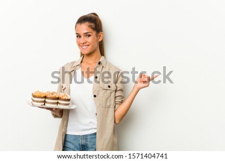 Young caucasian woman holding sweet cakes smiling and pointing aside, showing something at blank space.