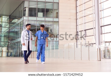 Male Medical Staff Talking As They Walk Through Lobby Of Modern Hospital Building Royalty-Free Stock Photo #1571429452