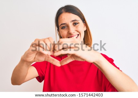 Beautiful redhead woman wearing casual red t-shirt over isolated background smiling in love doing heart symbol shape with hands. Romantic concept.