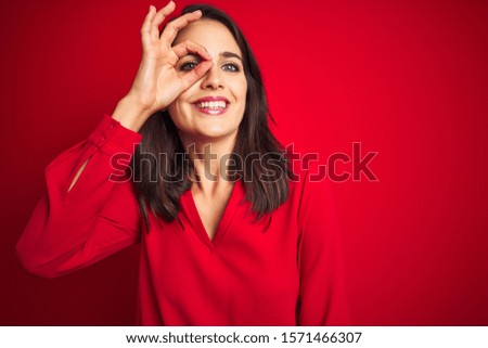 Young beautiful woman wearing shirt standing over red isolated background doing ok gesture with hand smiling, eye looking through fingers with happy face.