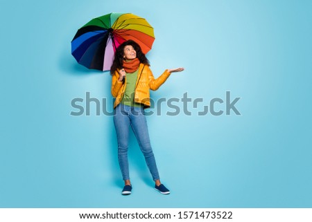 Full length photo of beautiful dark skin lady hold colorful umbrella catching rain drops enjoy amazing weather wear overcoat jeans green sweater scarf isolated blue color background