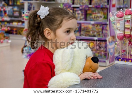 The girl is sad, holding a large polar bear and stands in the store among children's toys. Children's emotions, waiting for gifts and purchases.