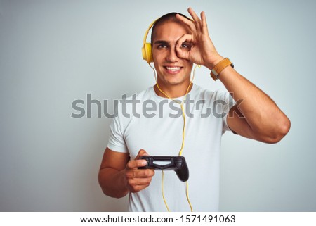 Young handsome man playing videogames using headphones over white isolated background with happy face smiling doing ok sign with hand on eye looking through fingers