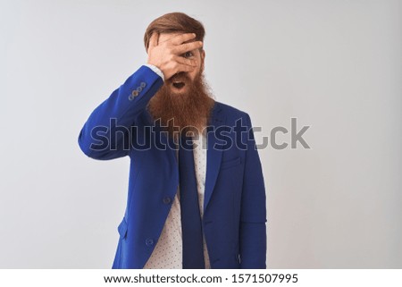 Young redhead irish businessman wearing suit standing over isolated white background peeking in shock covering face and eyes with hand, looking through fingers with embarrassed expression.