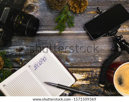 Photographer's plan for the New Year 2020. A camera, smartphone, tripod and notepad with a ballpoint pen lie on a wooden surface with green coniferous branches and cones and a cup of coffee.