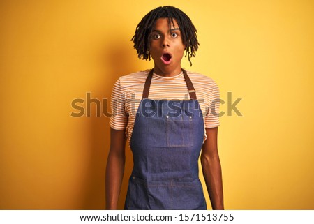 Afro american barista man with dreadlocks wearing apron over isolated yellow background afraid and shocked with surprise expression, fear and excited face.