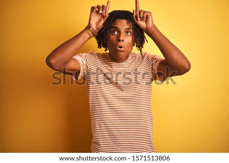 Afro man with dreadlocks wearing striped t-shirt standing over isolated yellow background doing funny gesture with finger over head as bull horns