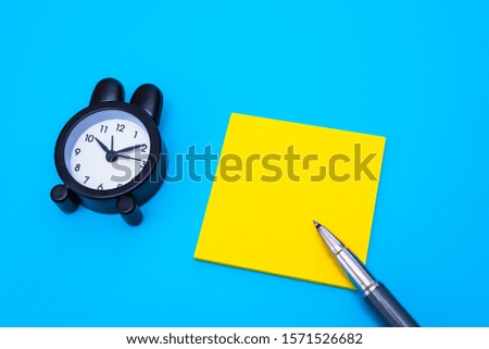 Flat lay of an alarm clock, pen and yellow paper to write any text, isolated on plain blue background.
