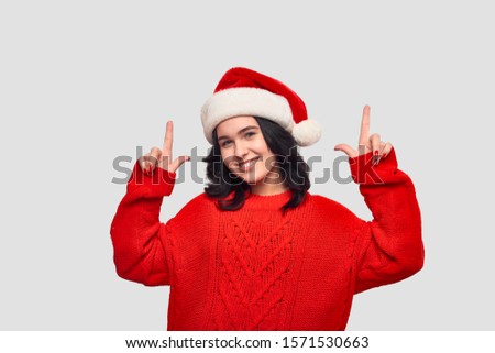 Pretty brunette girl in a red sweater and Santa hat pointing at the copy space up isolated over grey background. Celebrating Christmas and New Year