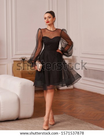 Full-body portrait of beautiful young woman with hair bun wearing black chiffon cocktail dress and high heels and posing in exquisite apartment. Magnificent brunette model in classy evening outfit. Royalty-Free Stock Photo #1571537878