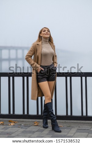 A long-haired attractive blonde walks along the autumn promenade in the morning. Fog. Blonde in a coat and boots. Posing for an autumn photo shoot
