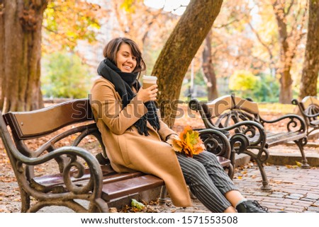 woman sitting on the bench at autumn city park drinking coffee