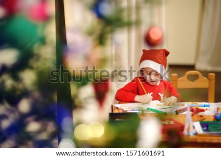 Little boy wearing in a Santa costume is drawing near a decorated Christmas tree. Leisure activities for children during the Christmas holidays at home.