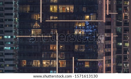 Rows of glowing windows with people in the interior of apartment buildings at night. Modern skyscrapers with glass surface. Concept for business and modern life. Zoom out