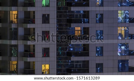 Rows of glowing windows with people in the interior of apartment building at night. Modern skyscraper from glass and concrete. Concept for business and modern life. Pan left
