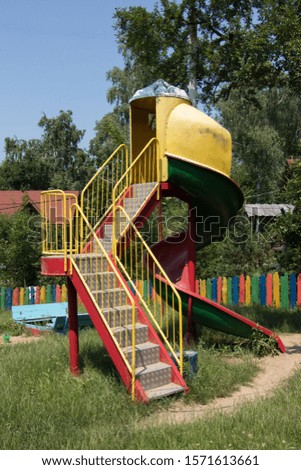 A vertical shot of a colorful playground slide with high trees and painted fence in the background
