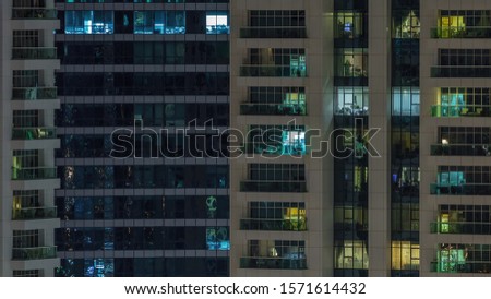 Rows of glowing windows with people in the interior of apartment building at night. Modern skyscraper with glass surface. Concept for business and modern life