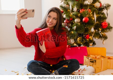 smiling excited pretty woman in red sweater sitting at home at Christmas tree unpacking presents and gift boxes taking selfie photo on phone camera