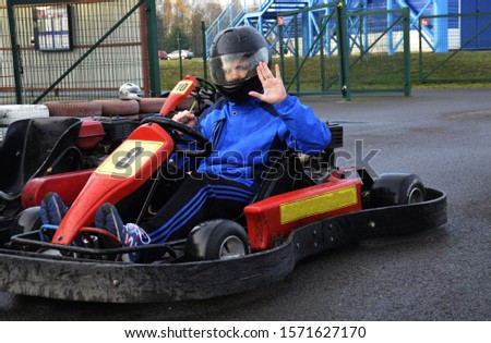 The guy in the black helmet is smiling, sitting in a race car, riding a go-kart. The young guy shows delight and waves to you. Sign: Hello and joy. He is in great anticipation of that high-speed ride.
