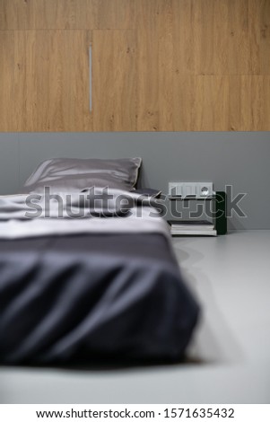 Amazing sleeping place with silk linens on the gray floor in the luminous interior with wooden walls. Next to it there are books, power socket with switches, thin lamp. Closeup. Vertical.
