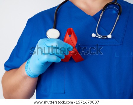 doctor in blue uniform and sterile latex gloves holds a red ribbon - a symbol of the fight against disease AIDS and vasculitis