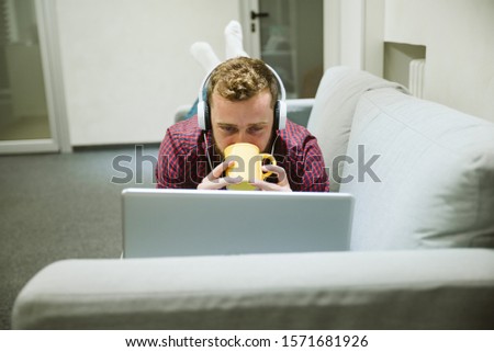 A bearded young man, lying on a bed, drinking coffee from a yellow cup and talking in a video call