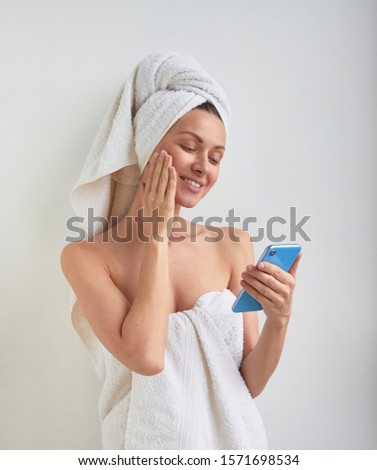 Glad beautiful young woman wrapped hair on head and body in spa bath shower towel touch skin on face and looks to the mobile phone screen selfie camera live translation in hand on white background Royalty-Free Stock Photo #1571698534
