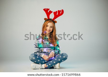 Beautiful little girl in a garland sitting widely smiles at the camera in the New Year. The red-haired lady wrapped a garland to create a festive mood. A teenager is sitting on the floor before