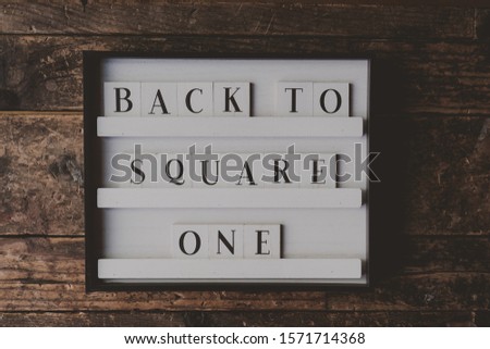 A white sign with writing "back to square one" on it on a wooden brown background