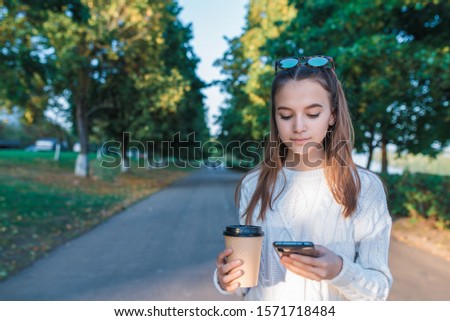 teenager girl in summer in city park, holding cup coffee tea, mobile phone, reads writes SMS, online application on social networks. Free space for copy text. Concept relaxation after school college