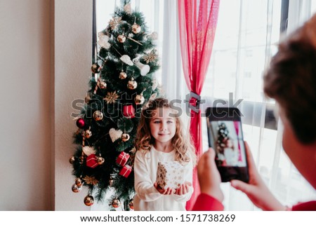 brother taking a picture of his sister with mobile phone at home. Girl standing by the christmas tree. Family time. Christmas concept