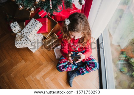 portrait of beautiful kid girl at home by the christmas tree and using mobile phone