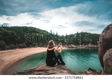 Young girl sitting on the rock with beautiful view on the beach and sea and looking into her phone
