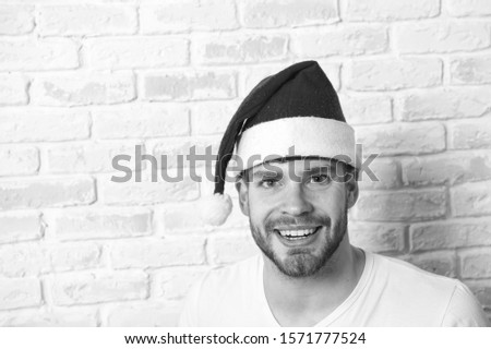 Man santa smile on white brick wall. Macho with bearded face in red xmas hat. Christmas and new year party. Santa claus fashion. Happy holidays celebration concept, copy space