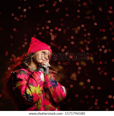 tle girl on a dark background in warm winter clothes, folded her hands in prayer, making a wish, looking up, on a dark background. Bokeh on background