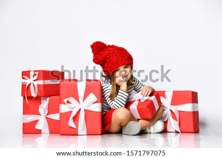 Portrait of a bored cute little girl in a red hat sitting in a pile of gift boxes, after shopping or after the holiday.