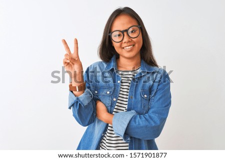 Young chinese woman wearing denim shirt and glasses over isolated white background smiling with happy face winking at the camera doing victory sign. Number two.